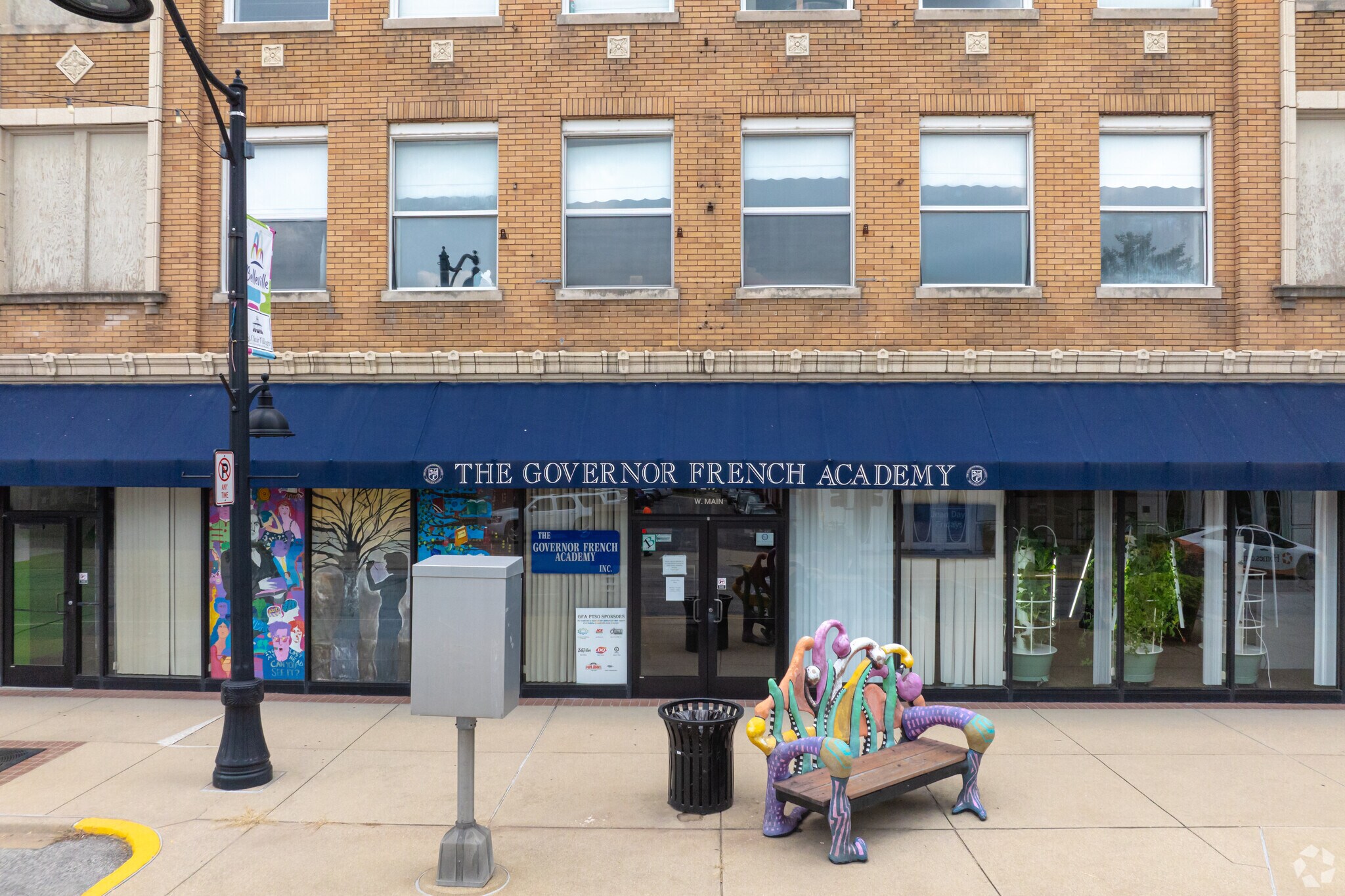 The Governor French Academy building exterior, featuring a blue awning, large windows, and a colorful, whimsical bench on the sidewalk.